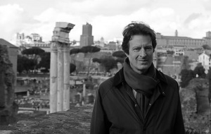 Black and white photo of man in front of old monuments wearing a scarf and coat with windswept hair