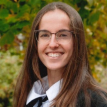 Naroa is standing in front of some trees. She is wearing sub fusc and glasses. She has long brown hair and is smiling.