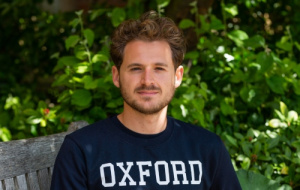 Ethan is sat on a wooden bench with foliage in the background. He is looking at the camera with a stoic expression, his hair is short, flowing, blonde curls and he has a cropped beard and is wearing a dark blue jumper.
