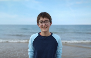 A head and shoulders photo of Rebecca smiling standing on a beach. They are wearing a navy sweatshirt with pale blue sleeves and glasses and have short brown hair. You can see the sea and end of the beach in the background.