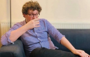 A picture of Tom sitting on a navy couch drinking a cocktail. He is wearing a purple and white chequered shirt and black jeans and has glasses and curly brown hair.