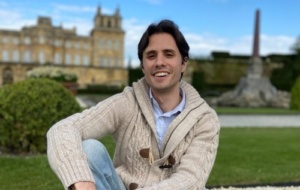 Carlos smiling and relaxed wearing a beige cable-knit jumper outside a castle