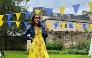 Gariyasi smiling wearing a yellow dress and blue jacket with Univ blue and yellow bunting in garden