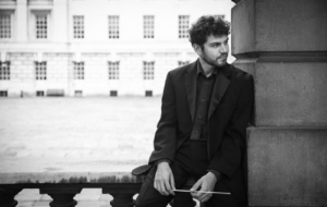 Man with curly dark hair with conductor's baton looking thoughtful in Oxford college