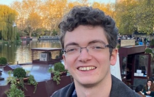 Man with short curly hair and glasses standing in front of river 