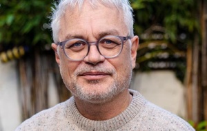 Middle-aged man with square frame glasses smiling looking at the camera wearing a beige round-necked jumper