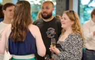 a man with a beard talks to two women, one holds a glass of wine