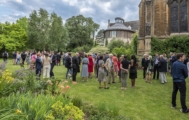A large group of smart-casually dressed people, many holding glasses, socialising in a garden