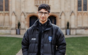 Young man with short black hair and square glasses standing on main quad in puffer with Univ crest and initials 
