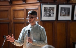 Young man with short black hair and square glasses wearing shirt and light grey jumper giving a speech