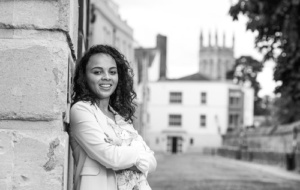 Woman with curly dark hair wearing white jacket leaning against Oxford wall, smiling at the camera 