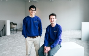 Two men in blue branded sweatshirts white background on sits on a box, the other stands with his hands in his pockets