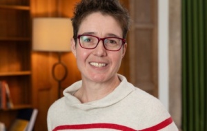 Woman with short brown hair wearing red glasses and a white and red striped jumper smiling at the camera