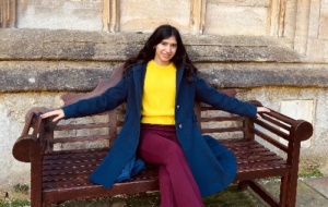 Woman sitting on a bench wearing yellow jumper, blue coat, and burgundy trousers