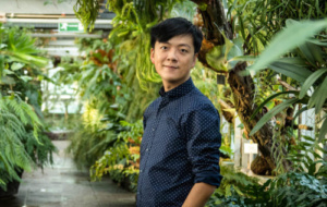 Man with dark hair wearing a blue shirt standing in a botanical garden looking at the camera
