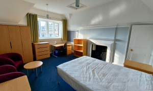 A bedroom for an undergraduate student. There is a single bed with bedside table, desk and office chair, and two sitting chairs with a small table. There is a large window and wardrobe, and an old-fashioned fireplace.